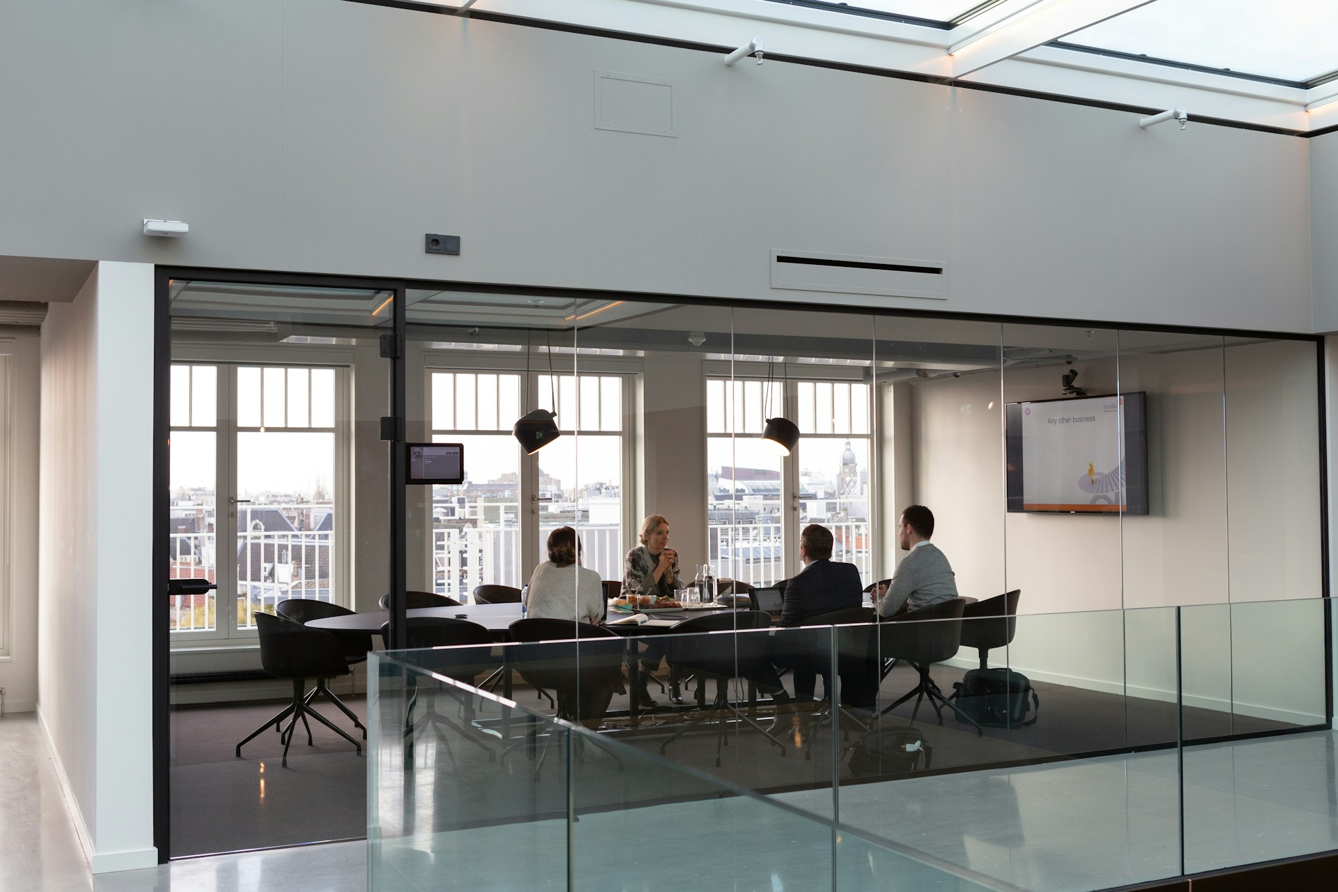 Four people in a modern glass-walled meeting room engaged in discussion around a conference table, with a cityscape visible through large windows.