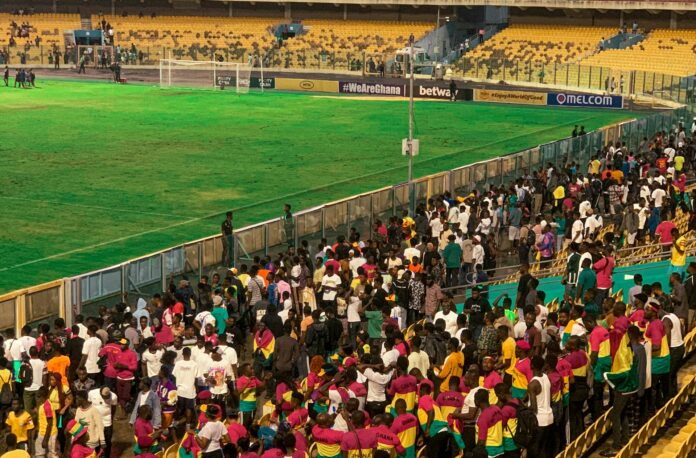 A crowd of Ghanaian football fans gathers in the stands of Accra Sports Stadium, wearing colorful national team jerseys, as they wait near the pitch after a match.
