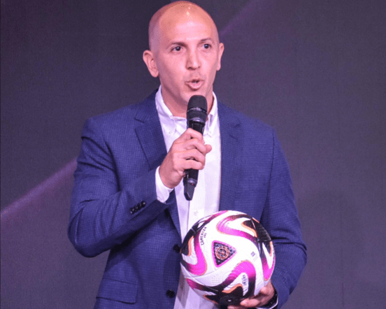 Vicente Suanno speaking on stage in a blue blazer, holding a microphone and a pink-and-black football