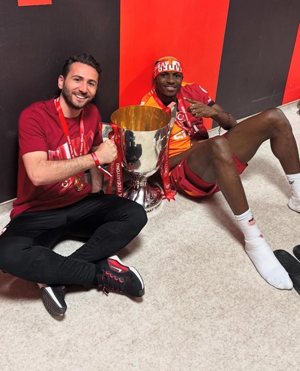 Vural Durmuş celebrating with a Galatasaray player in the dressing room, sitting beside the championship trophy.