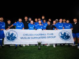 Chelsea Muslim Supporters Group team holding banner on pitch after a match under floodlights.
