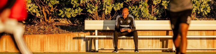 Andrea Canna observes training attentively while seated on the sidelines during a sunlit session.